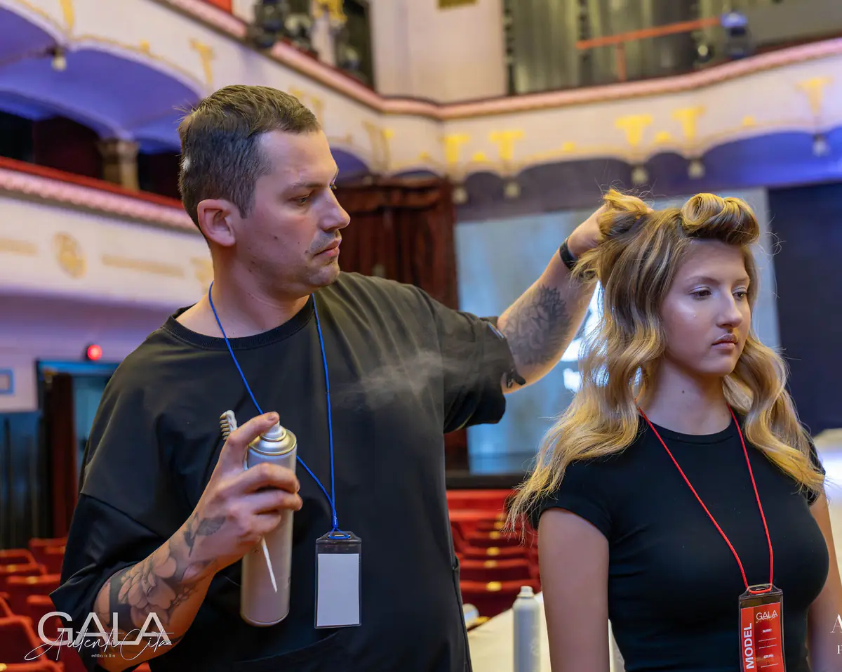 Attila setting a model's waves with hairspray on a theatre stage during a gala